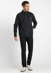 Veste noire avec des accents de zip bleu, fermeture éclair à l'avant, col montant et coupe slim. Assortie avec un pantalon noir et des baskets blanches.