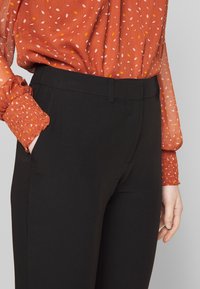 Sorte bukser med en glat tekstur og skræddersyet pasform, der har sidelommer. Kombinert med en teksturert oransje bluse med et spraglete mønster.