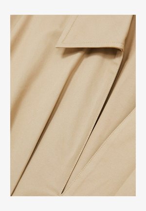 Beige stof, gladde textuur, met een kraag en subtiele plooien. Close-up weergave die stikseldetails en natuurlijke plooien benadrukt.