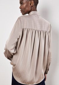 Blusa di seta in morbido beige con righe verticali e design con schiena arricciata. Presenta maniche lunghe con polsini bottonati e colletto classico.