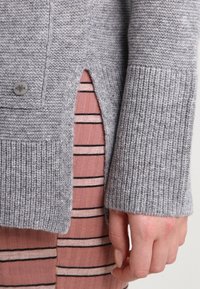 Gray knitted sweater with ribbed cuffs and side slit, featuring a button detail. Underneath, a pink striped fabric is partially visible.
