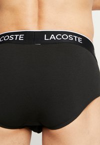 Fekete pamut alsónadrág, márkás elasztikus derékpánttal, amelyen fehér színnel van nyomtatva a "LACOSTE" felirat. Sima textúra és testhezálló kialakítás.