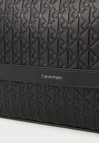 Fekete textúrájú bőr táska geometrikus mintával és domború motívumokkal. Egy elegáns fekete pánt található rajta, amelyen ezüst színű "Calvin Klein" logó van.