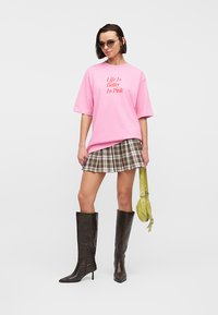 Γυναίκα που φοράει ροζ φαρδιά μπλούζα με την επιγραφή "Η ζωή είναι καλύτερη στο ροζ", καρό μίνι φούστα, μαύρες μπότες μέχρι το γόνατο, γυαλιά ηλίου, κρατώντας μια πράσινη τσάντα.