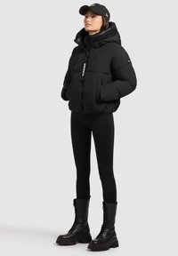 Juodas pūkinis švarkas su kapišonu, su priekyje esančia užtrauktuku, dekoratyvinėmis žymėmis ir dviem priekinėmis kišenėmis, derinamas su juodom kelnėm ir kariškais batais.