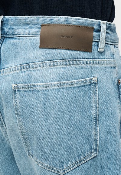 Ανοιχτό μπλε τζιν με ομαλή υφή, που διαθέτει σκούρο καφέ δερμάτινο έμπλαστρο στη πίσω μέση και δύο πίσω τσέπες.