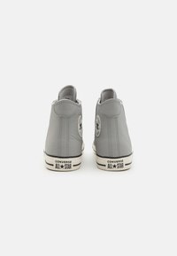 Adidași înalți din piele gri, cu un patch logo rotund, talpă din cauciuc alb contrastant și accente negre, având textul "CONVERSE ALL STAR".