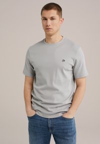 Heled hall puuvillane T-särk ümmarguse kaelusega, lühikeste varrukatega ja väikese musta logoga vasakul rinnal. Seljas on sinised teksapüksid.