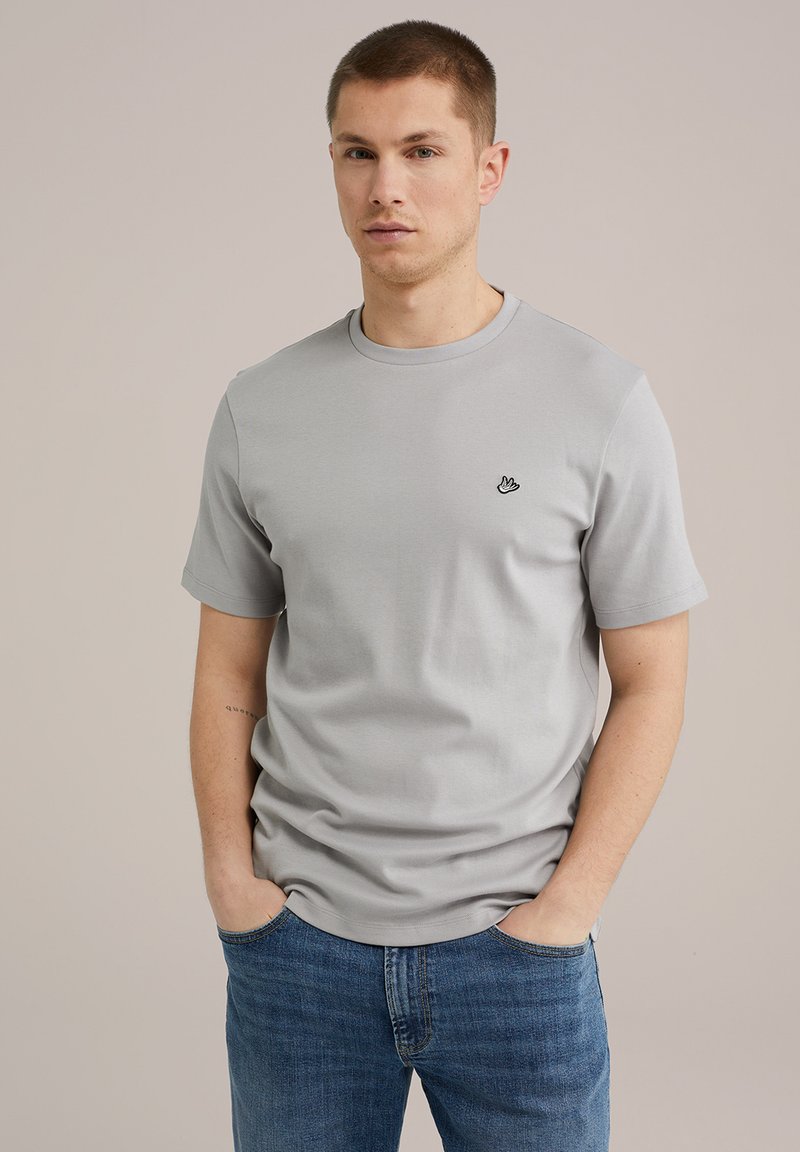 Heled hall puuvillane T-särk ümmarguse kaelusega, lühikeste varrukatega ja väikese musta logoga vasakul rinnal. Seljas on sinised teksapüksid.