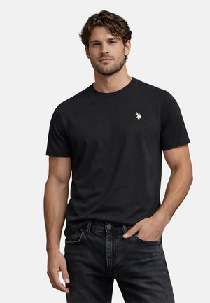 Mann mit lockigem Haar, der ein schwarzes T-Shirt mit weißem Polo-Player-Logo und schwarze Jeans trägt, steht mit einer Hand in der Tasche.