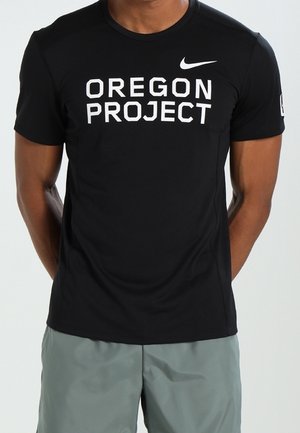 Hombre con camiseta negra de Nike con el texto "OREGON PROJECT", combinado con pantalones cortos grises, posando frente a un fondo liso.