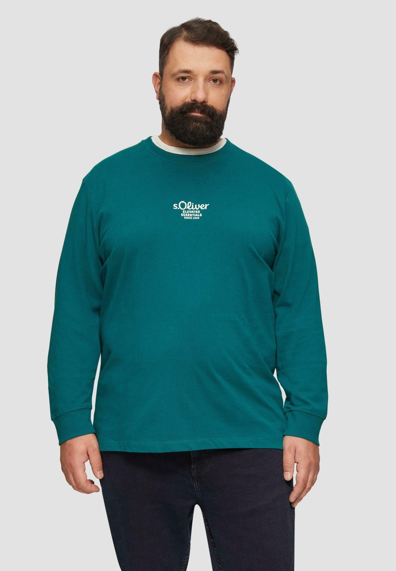 Langärmliges T-Shirt in Petrol mit weißem gesticktem Logo, Rundhalsausschnitt und lockerer Passform. Getragen mit dunklen Jeans.