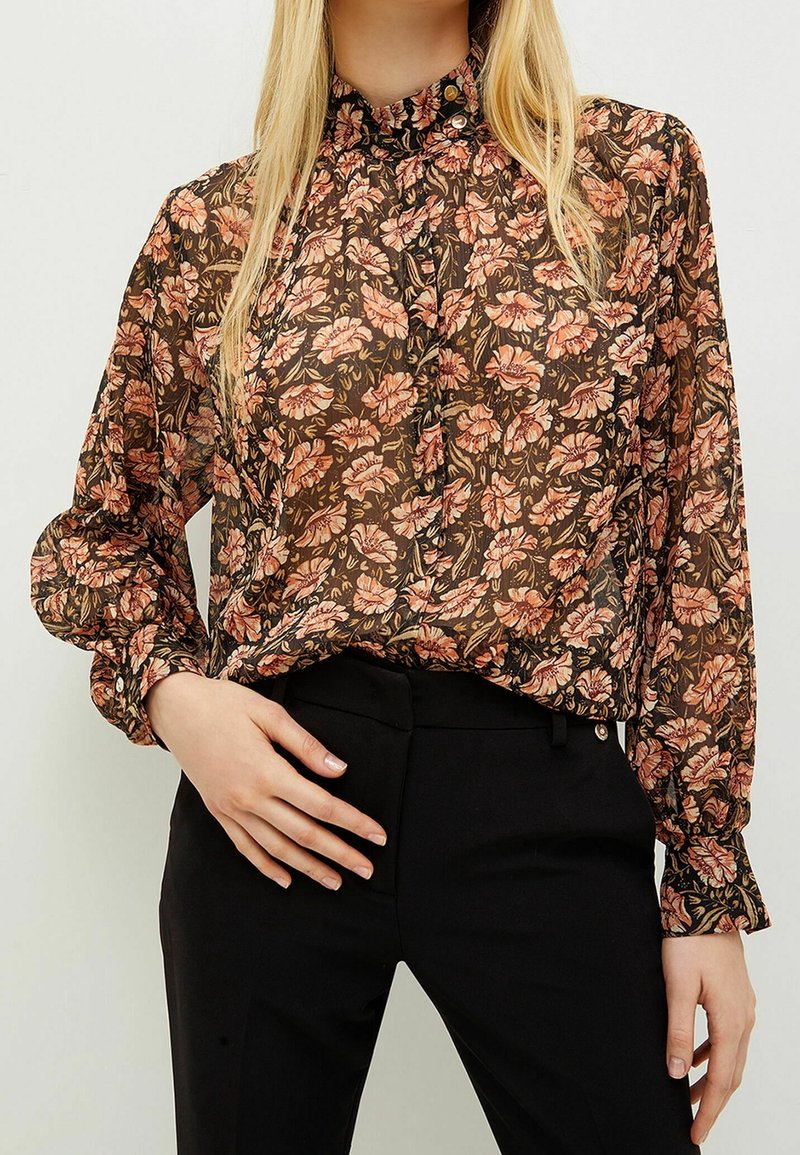 Femme portant un chemisier fleuri transparent à manches longues avec des fleurs roses et marron, rentré dans un pantalon noir taille haute.