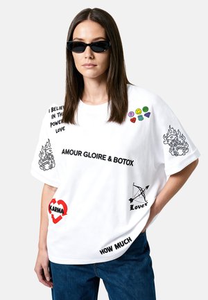 Femme portant des lunettes de soleil noires et un t-shirt blanc oversize avec différentes phrases imprimées et des patches graphiques colorés.