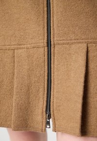 Gros plan d'une jupe en laine marron avec une fermeture éclair verticale noire et des plis près du bord inférieur montrant une partie des jambes nues.