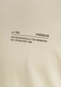 Kremno obarvana majica s kratkimi rokavi z natisnjenim rjavim besedilom. Besedilo vključuje "JJ-1990," "PREMIUM," in informacije o ustanovitvi leta 1990.