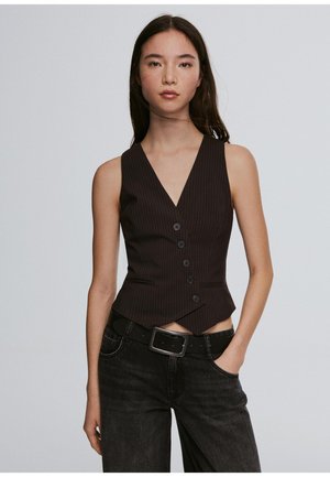 Jonge vrouw met lang donker haar draagt een nauwsluitend zwart gilet met krijtstreepknopen, zwarte riem en donkere spijkerbroek, staand met één hand in haar zak.