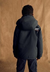 Giacca blu scuro con cappuccio, realizzata in tessuto testurizzato, con una toppa del logo sulla spalla sinistra e polsini elasticizzati. Indossata con pantaloni abbinati.