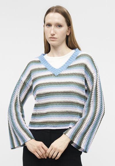 Femme portant un pull ample à rayures tricotées avec des tons bleus, verts, blancs et violets, associé à un sous-pull blanc et un pantalon noir.