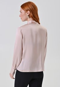 Helle rosa Satin-Bluse mit langen Ärmeln und hohem Kragen, mit geknöpften Manschetten und glatter Textur, getragen mit schwarzen Hosen.