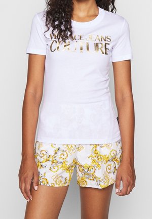 Vrouw met een wit T-shirt met korte mouwen en een gouden "Versace Jeans Couture" logo, en een witte korte broek met een ingewikkeld geel-zwart barokpatroon.