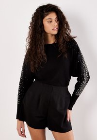 Schwarzer, gestrickter Pullover mit langen Ärmeln, verziert mit weißen Perlen; kombiniert mit hochgeschnittenen schwarzen Shorts, die ein maßgeschneidertes Design aufweisen.