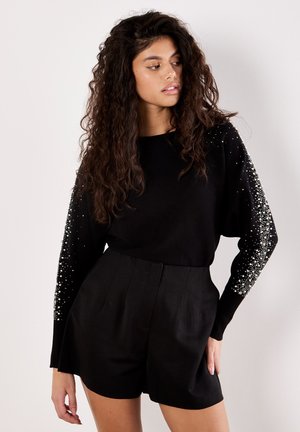 Maglione nero lavorato a maglia con maniche lunghe impreziosito da perline bianche; abbinato a pantaloni corti neri a vita alta, con un design su misura.
