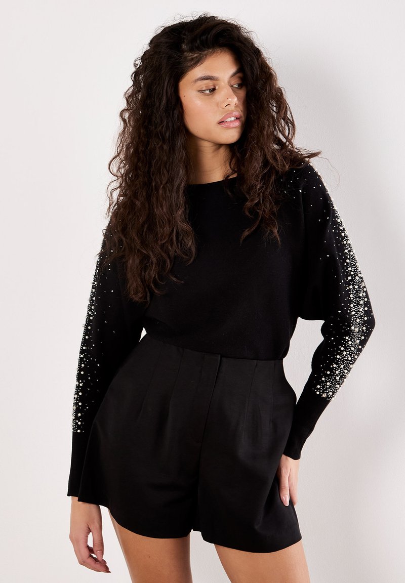 Schwarzer, gestrickter Pullover mit langen Ärmeln, verziert mit weißen Perlen; kombiniert mit hochgeschnittenen schwarzen Shorts, die ein maßgeschneidertes Design aufweisen.