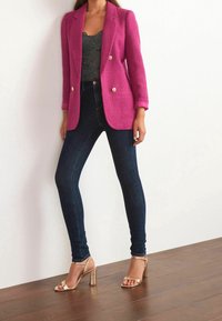 Blazer texturé magenta avec boutons dorés, associé à un jean ajusté bleu foncé et des sandales à talons hauts dorées à brides sur un sol en bois.