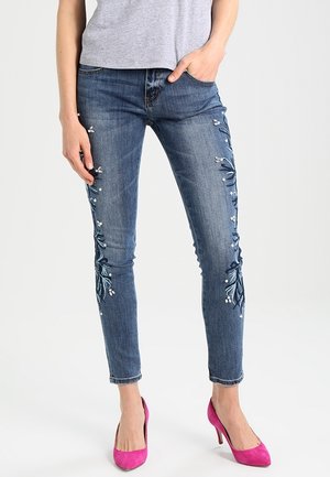 Jean en denim bleu à coupe slim avec broderies florales bleues et blanches sur les côtés, associés à des talons roses vifs.