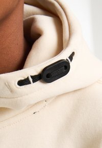 Felpa leggera beige con una consistenza morbida, dotata di un cordino nero e di un toggle regolabile al collo. Dettaglio ravvicinato del tessuto.