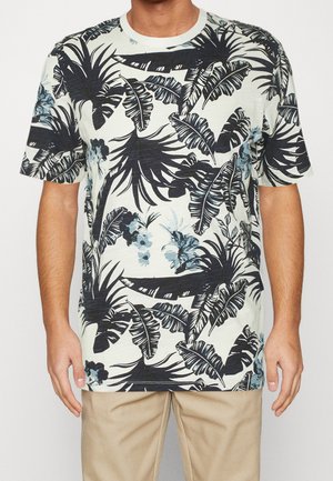 Homme portant un t-shirt blanc à manches courtes avec un imprimé de feuilles tropicales et de fleurs noires et bleues, associé à un pantalon beige.