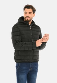Uomo che regola le maniche di una giacca nera imbottita con cappuccio, indossando jeans blu, in piedi davanti a uno sfondo bianco uniforme.