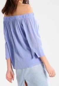 Blouse lavande décolletée avec des manches bouffantes, un col élastique et des poignets fendus, fabriquée en tissu lisse. Portée sur une jupe en denim légère.