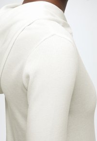 Közeli felvétel egy fehér bordázott hosszú ujjú felsőt viselő személyről, váll- és felkar részletek láthatók egy egyszínű háttér előtt.