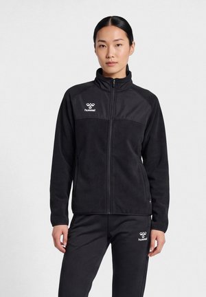 Млада жена с черна коса носи черно яке с цип Hummel и подходящи панталони, стои на бял фон.