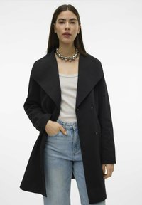 Svart oversized kappa med bred krage, dubbelknäppt design och textilerat tyg. Bärs över en ljus topp och blåjeans.