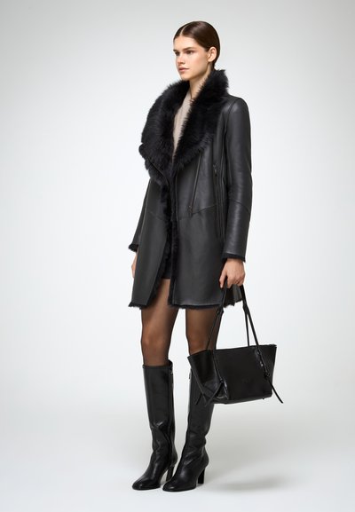 Schwarzer Ledermantel mit einem Fellkragen, knielange schwarze Lederstiefel und eine schwarze Handtasche. Glatte Textur mit Reißverschlüssen und strukturiertem Design.