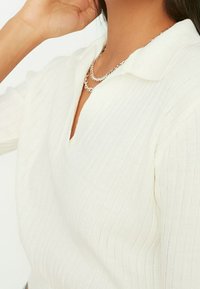 Weißes, geripptes Strickoberteil mit Notchkragen, kurzen Ärmeln und einer subtilen Textur. Das Outfit wird durch eine geschichtete silberne Kettenhalskette ergänzt.