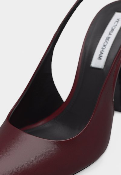 Temno rdeč usnjen čevelj z visokim podplatom in paščkom za peto, s koničasto konico in črto notranjo oblogo, z belo oznako oblikovalca na podplatu.