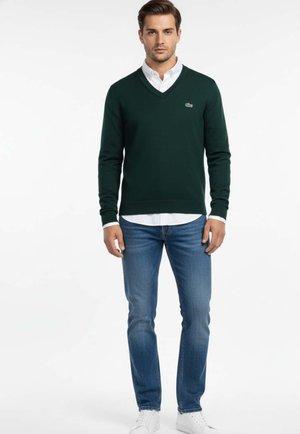 Jersey verde de cuello en V hecho de material de punto, combinado con una camisa blanca abotonada y jeans azules. Logo de marca destacado en el pecho izquierdo.