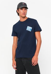 Sötétkék pamut póló, rövid ujjakkal és kerek nyakkivágással. Bal mellen fehér és kék "Tommy Jeans" grafika látható.