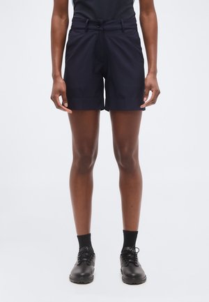 Persona, kas valkā melnas šūtas šortus, melnas zeķes un melnas mežģīnēs kurpes, stāvot pret tīru balto fonu.