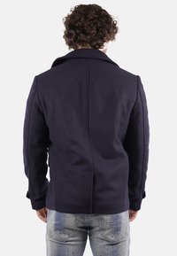 Cappotto blu navy in misto lana con colletto e tasche laterali, caratterizzato da un design posteriore sartoriale e dettagli in contrasto nelle cuciture. Vestibilità casual.