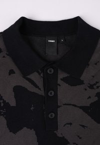 Svart polo-skjorte med teksturert overflate, med et kamuflasjemønster i mørkegrått. Den har en flat krage og tre svarte knapper.