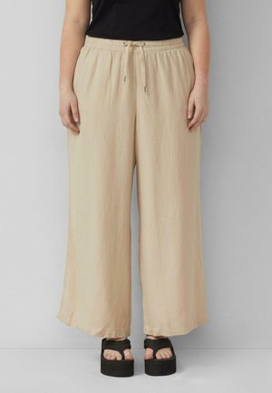 Person trägt beige weite Hosen mit Tunnelzug in der Taille und schwarze Plateau-Sandalen, stehend vor einem einfarbigen hellgrauen Hintergrund.