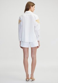 Camicia bianca con accenti ricamati in oro sulle maniche, caratterizzata da maniche lunghe, polsini con bottoni e un orlo curvo, abbinata a pantaloni corti bianchi.