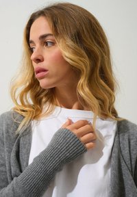 Cardigan grigio a coste indossato sopra una maglietta bianca, con tessuto morbido e maniche lunghe con polsini aderenti. Capelli acconciati in morbide onde.