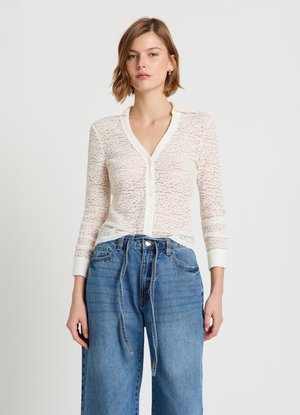 Spitzenstrickjacke in Off-White mit V-Ausschnitt, Knopfverschluss und langen Ärmeln. Kombiniert mit hoch taillierten, weiten Jeans aus blauem Denim.