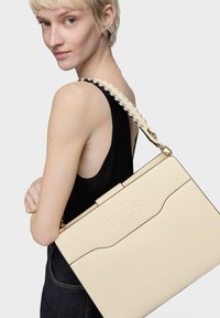 Borsa tote in pelle beige con finitura testurizzata e dettagli in oro. Presenta una tracolla intrecciata e il logo "TOUS" in rilievo sulla parte frontale.